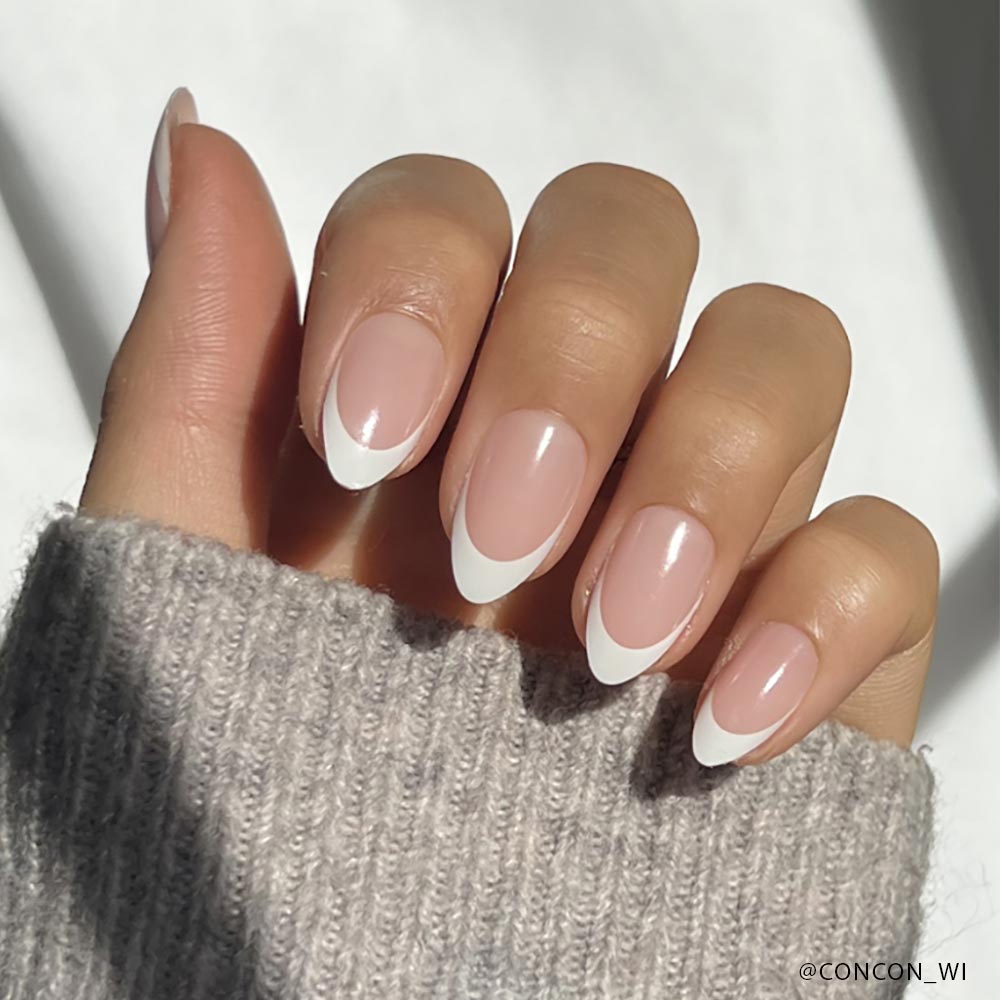 A hand with almond-shaped Ma Damn nails by glamnetic, showcasing a classic French manicure with a pale pink base and white tips, rests against a gray knitted sleeve.